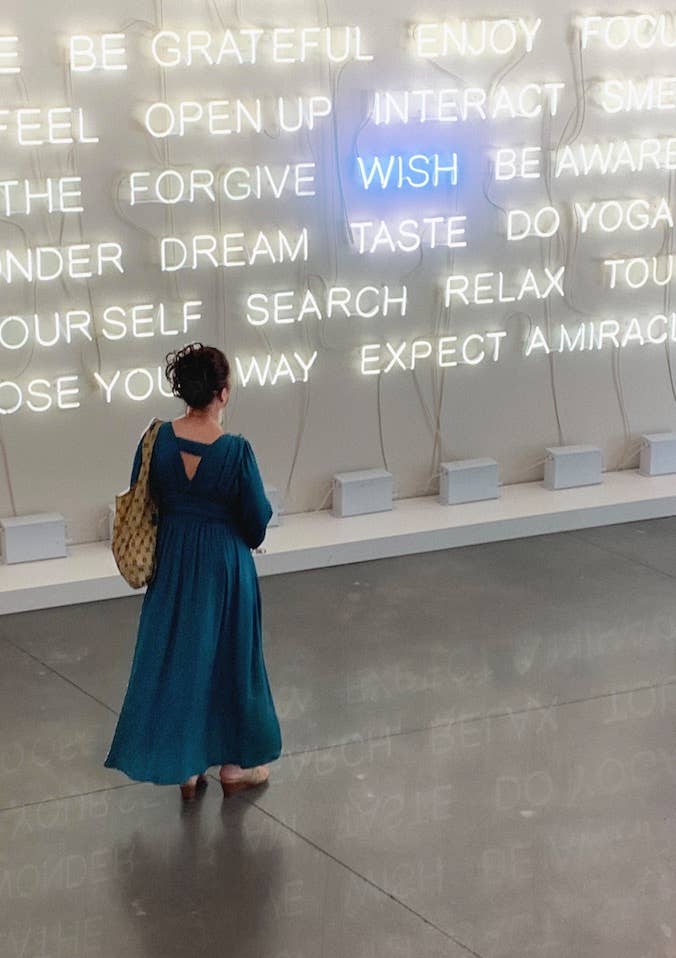 Woman standing in front of illuminated inspirational words featuring 'WISH' in blue at a contemporary art exhibit.