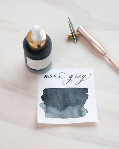 Moon Grey Calligraphy Ink with gold label and pipette, displayed with a calligraphy pen and ink swatch, available at stationery store.