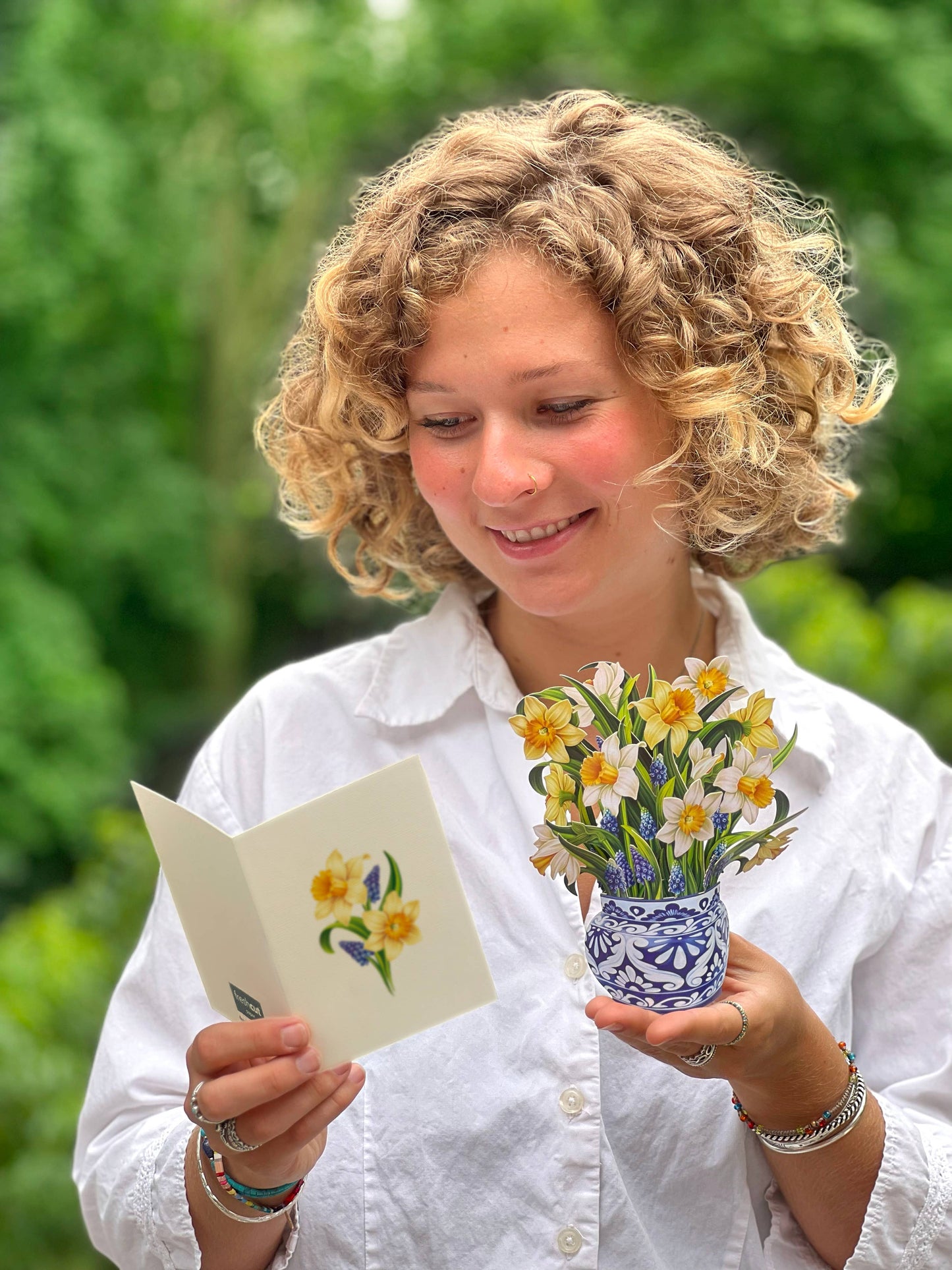 Woman holding Mini English Daffodil bouquet and note card from a stationery store, set against a green garden background.