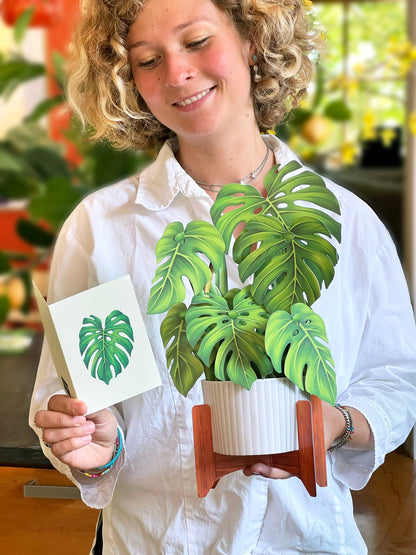 Person holding Monstera Plant Pop-up Greeting Card with mahogany stand and note card at a stationery store.