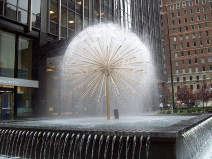 Large dandelion fountain sculpture surrounded by water jets in urban plaza with modern and historical buildings in the background.