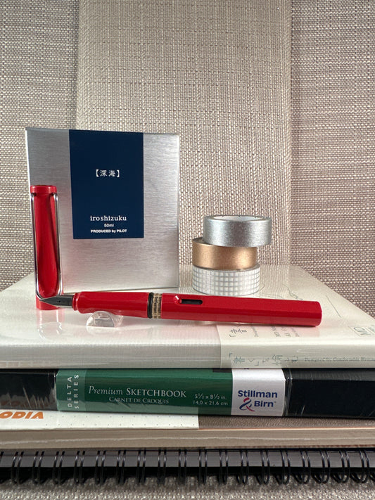 Red LAMY Safari Fountain Pen on sketchbook with ink and stationery supplies in background, ideal for a stationery store display.