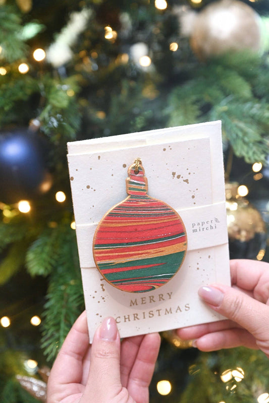 Festive Keepsake Marble Ornament Christmas Card with bauble design held in front of a decorated tree, available at a stationery store.