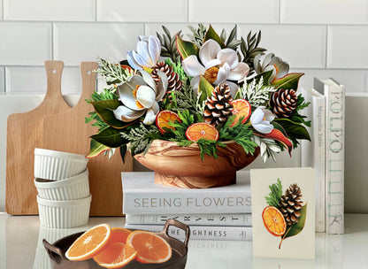 Winter Magnolia Pop-up Greeting Card with magnolias, orange slices, and pine cones in wooden bowl, perfect for stationery store display.