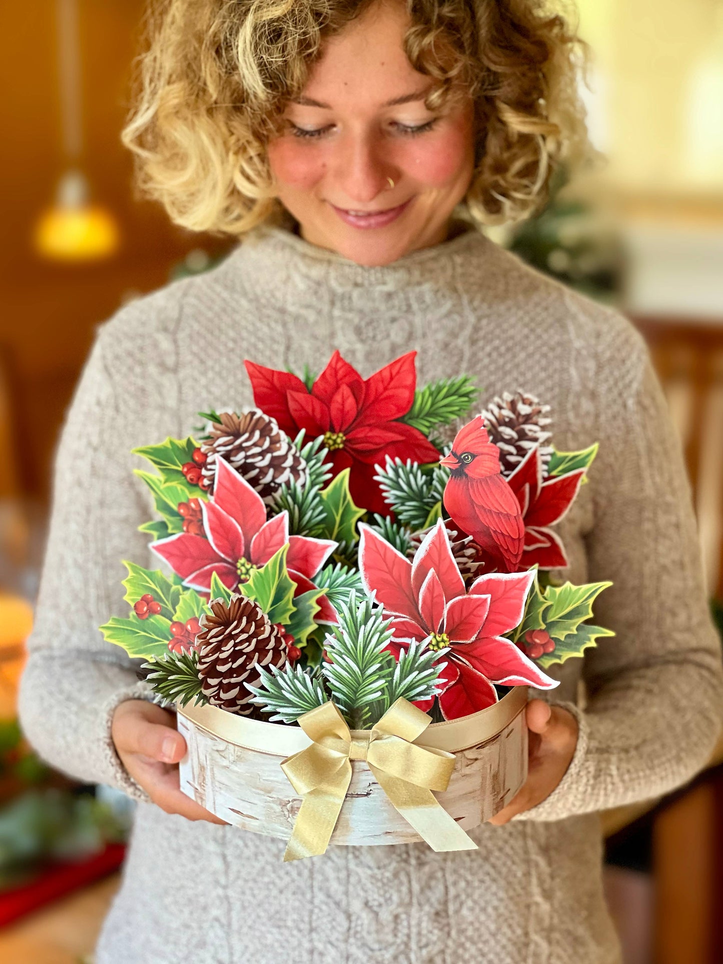 Woman holding Birch Poinsettia pop-up greeting card from stationery store, featuring pinecones, holly, red poinsettias, and champagne bow.